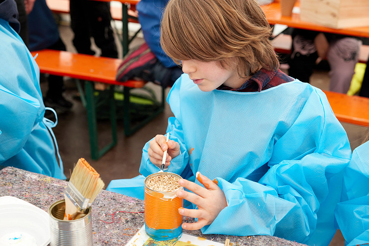 Ein Kind malt mit einem Pinsel eine Konservendose orange an. Die Dose ist gefüllt mit Bambusröhrchen.