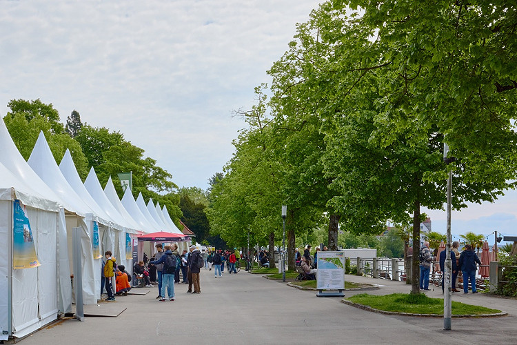 Eine breite Promenade am Wasser mit einer Reihe weißer Zelte auf der linken Seite, an denen Menschen stehen und sich informieren. Rechts säumen große grüne Bäume den Weg, einige Personen sitzen auf Bänken oder gehen spazieren. Im Hintergrund ist das Ufer mit Geländer und vereinzelten Pflanzen zu sehen.